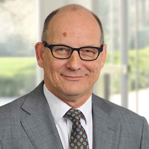 Man in a grey suit with a patterned tie, set against a bright office background.