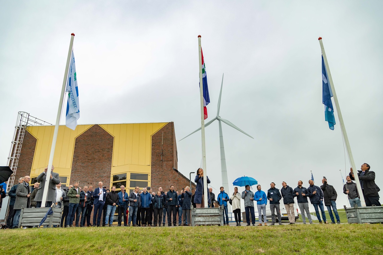 A gathering of people at a flag-raising event near a wind turbine and a building with a yellow roof.