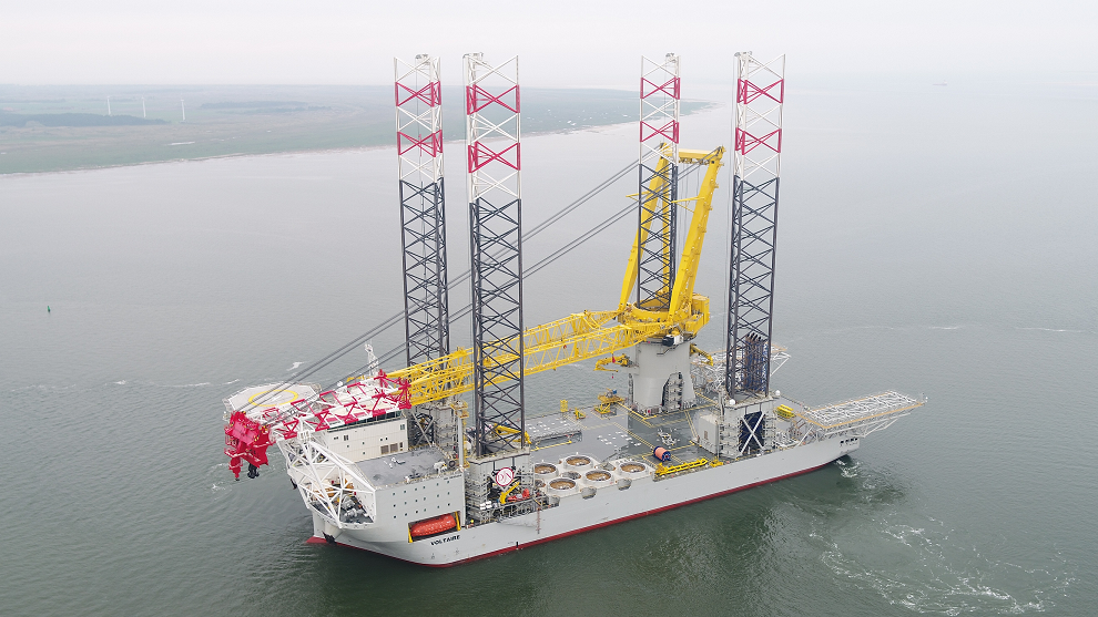 Een groot offshore constructieschip met gele en rode accenten werkt in kalme wateren, met windturbines op de achtergrond.