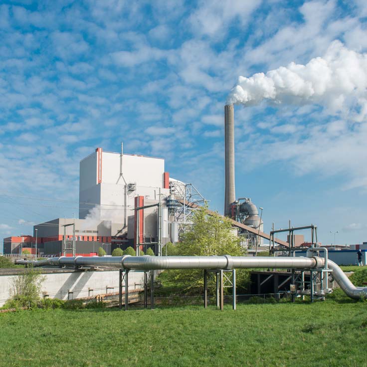 Een energiecentrale met rook die uit een hoge schoorsteen komt, omgeven door groen gras en industriële leidingen onder een blauwe lucht.