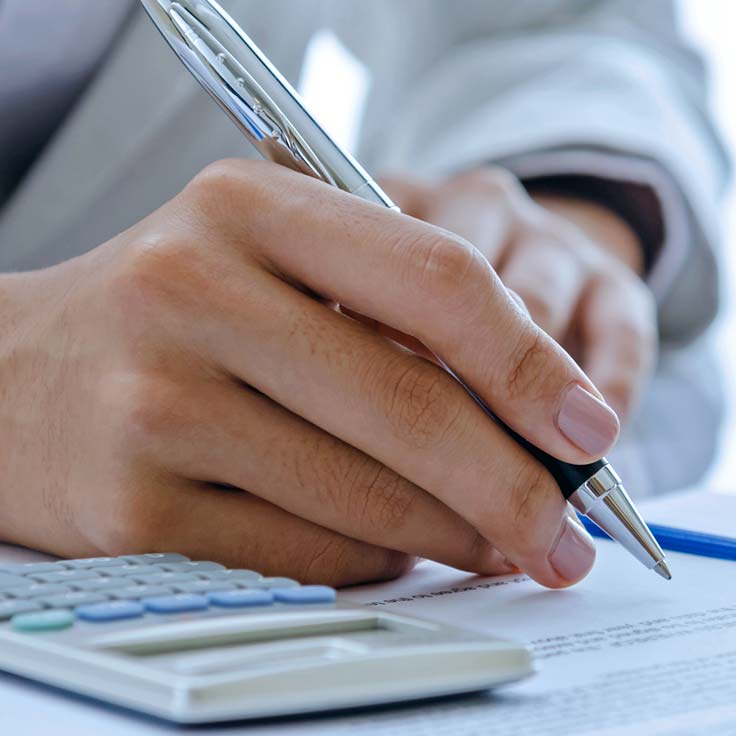 A close-up of a hand holding a pen, poised over documents with a calculator in the foreground.