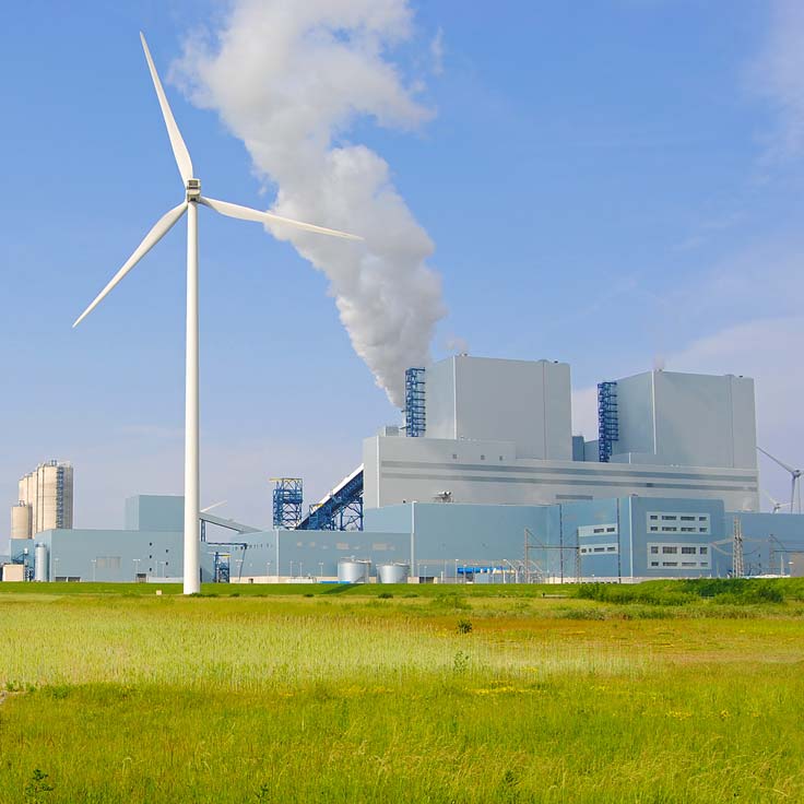 A wind turbine stands in the foreground with a power plant emitting smoke in the background under a clear blue sky.