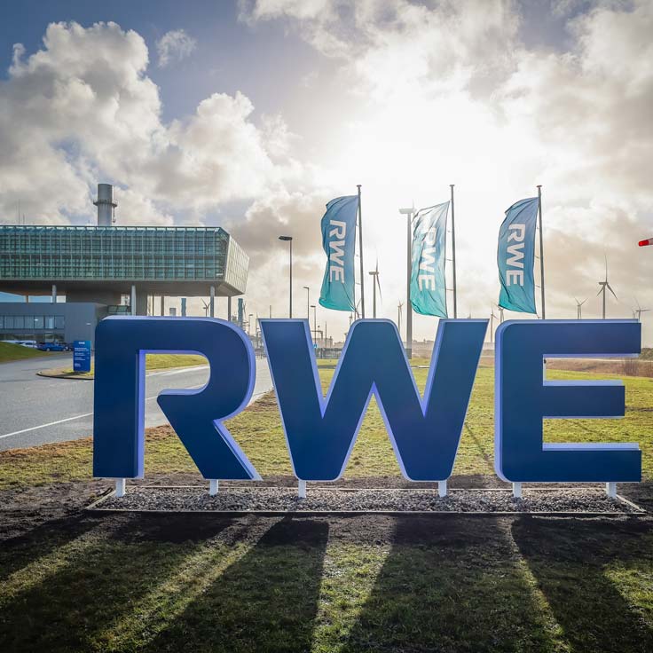 Het RWE-sign bij een kantoorgebouw, omringd door vlaggen en windturbines onder een bewolkte lucht.