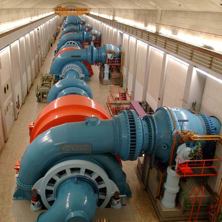 Aerial view of a power generation facility with multiple large turbines and pipes in a long hall, featuring blue and orange colours.