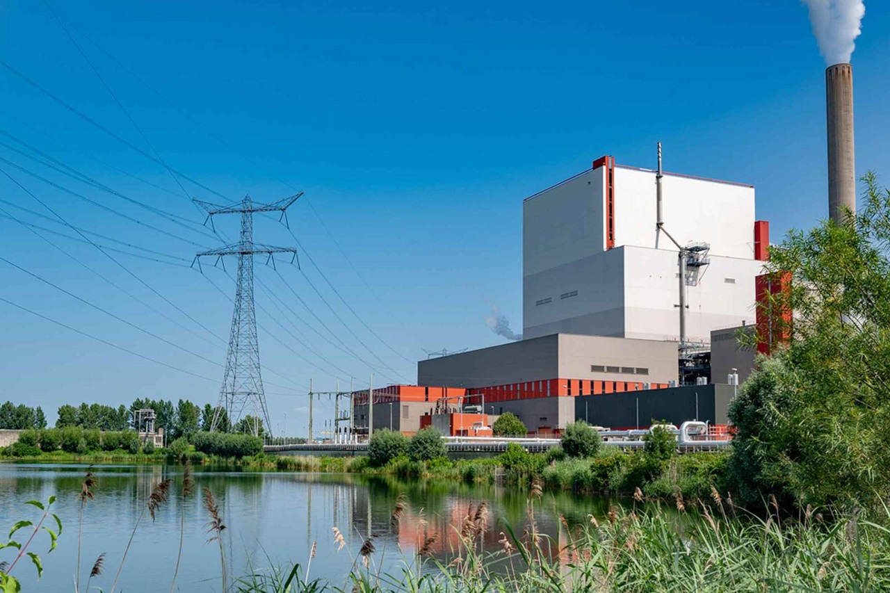 Een energiecentrale bij een rustig meer, met een hoge schoorsteen en elektriciteitskabels tegen een heldere blauwe lucht.