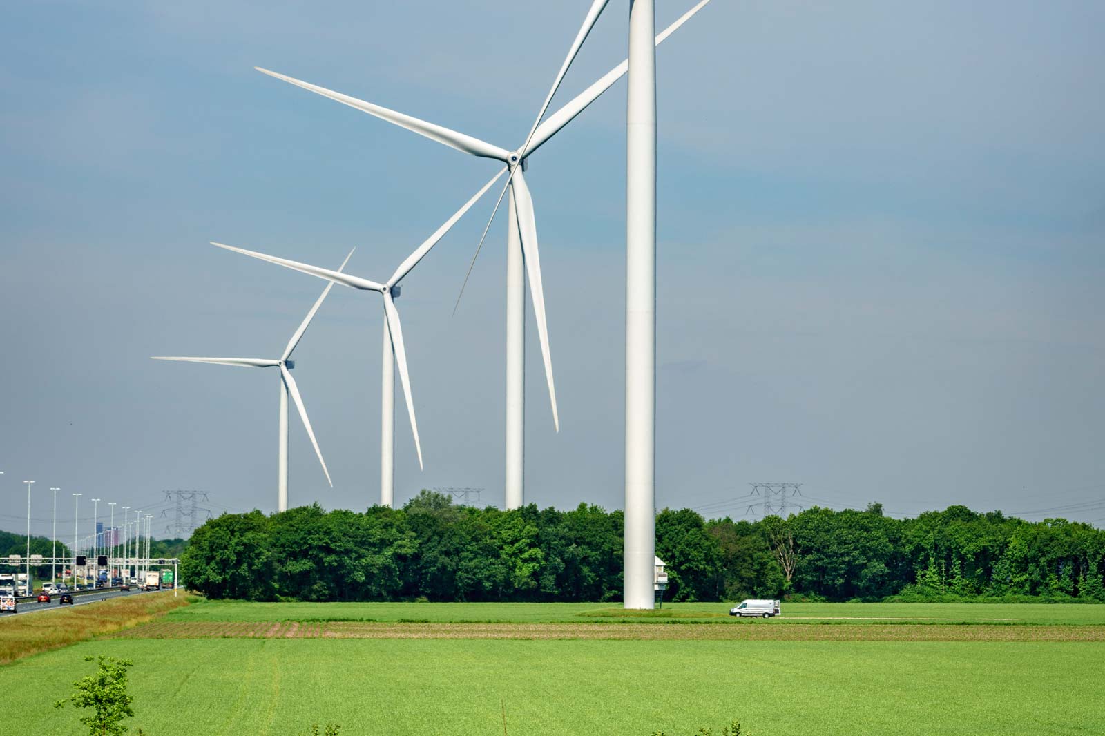 Wind turbines stand tall beside a green field and road. A van drives along the road under a clear blue sky.