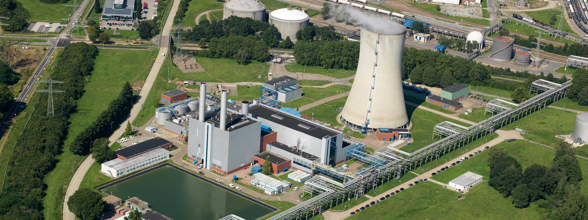 Luchtbeeld van een energiecentrale met een koeltoren, grote leidingen en een wateroppervlak op de voorgrond.