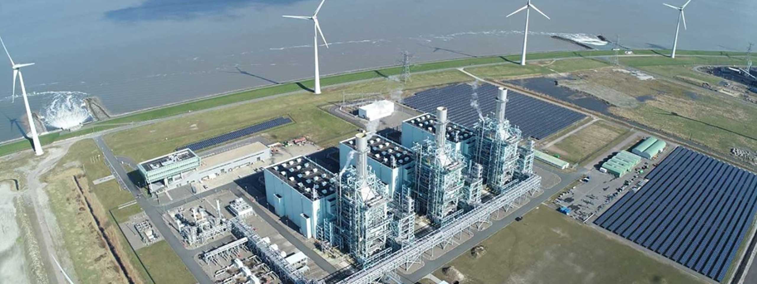 Een luchtfoto van een energiecentrale met windturbines en zonnepanelen, gelegen nabij een waterlichaam.
