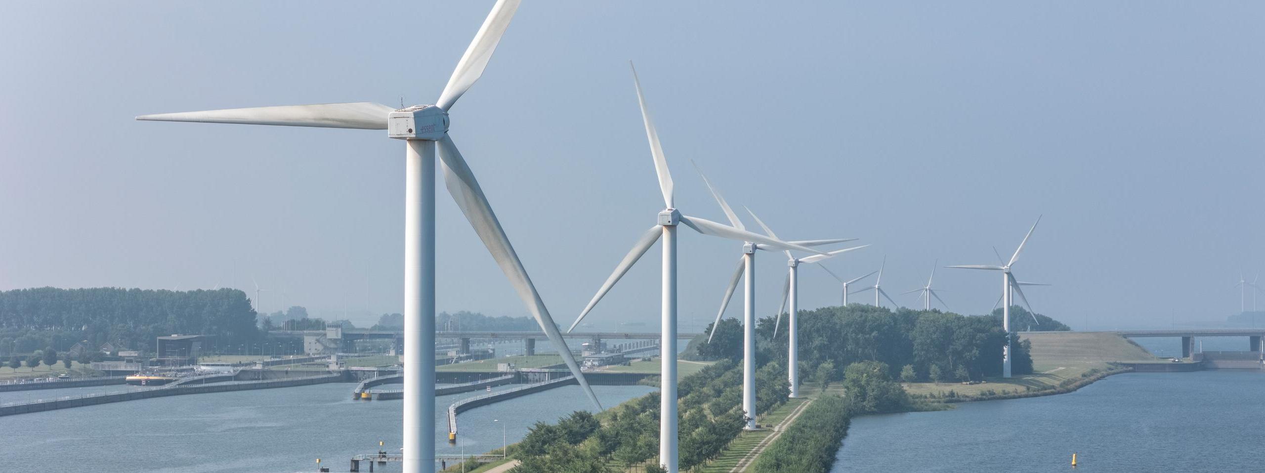 Een panoramisch uitzicht op meerdere windturbines naast een rivier met een brug op de achtergrond onder een heldere lucht.