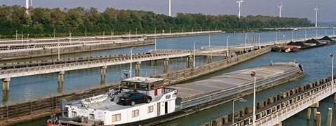 Een schip op een rivier, met windturbines op de achtergrond en industriële kades langs de oever.
