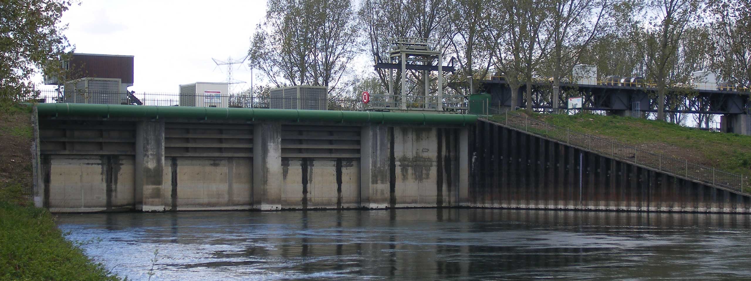 Een hydraulische structuur met een groene buis bovenop, met uitzicht op een rivier, omringd door bomen en gebouwen.