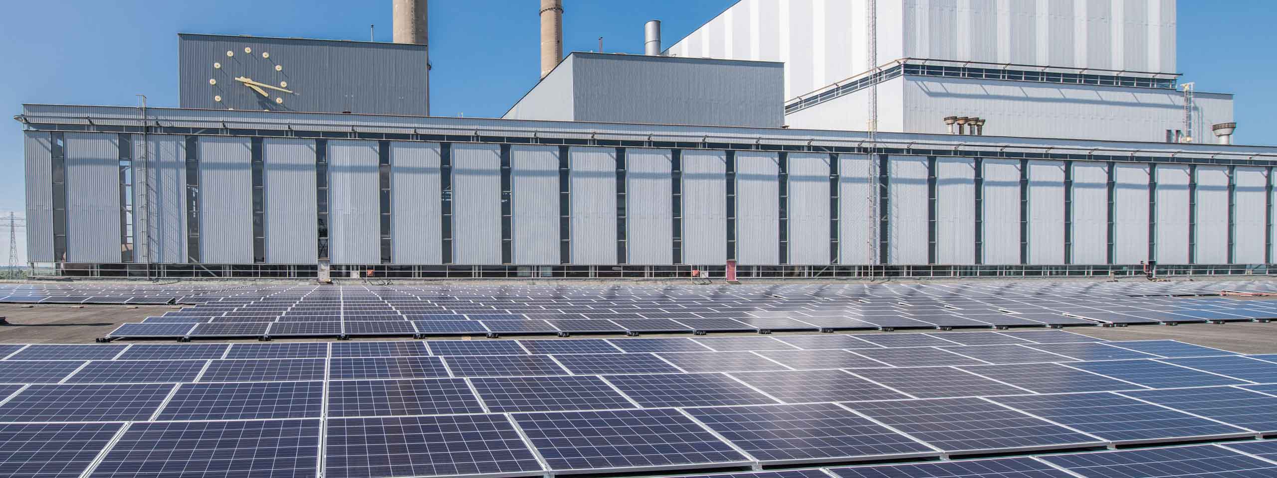 Zonnepanelen bedekken het dak van een groot industrieel gebouw, met een klok en schoorstenen zichtbaar tegen een heldere blauwe lucht.