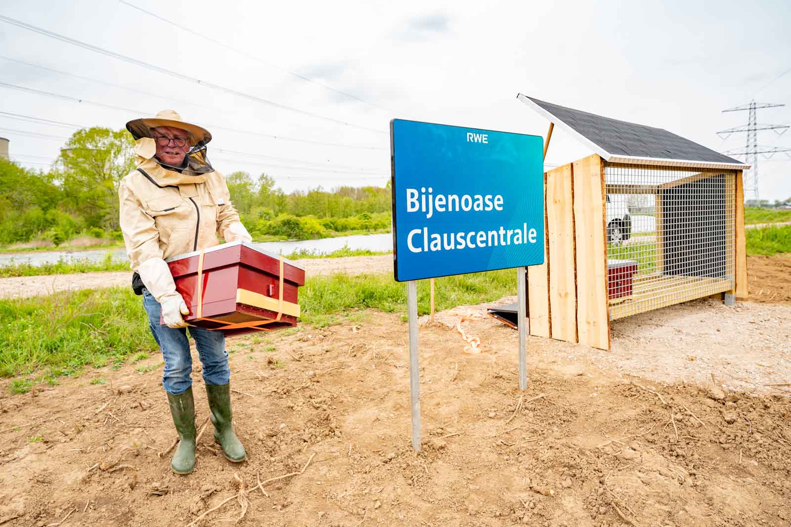 A person holding a bee box stands in front of a sign that reads 'Bijenoase Clauscentrale'.