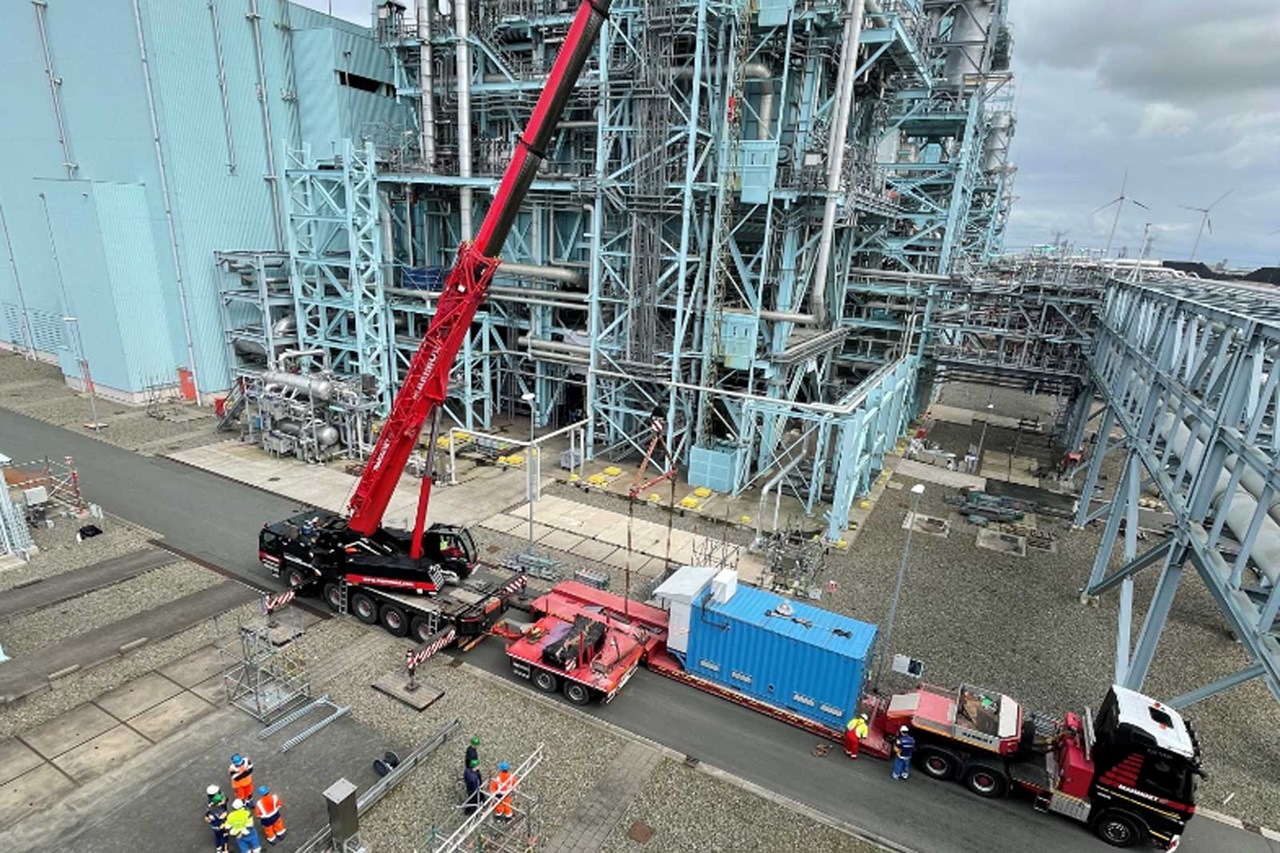 A red crane lifts equipment near a large industrial facility with workers overseeing operations on a gravel path.