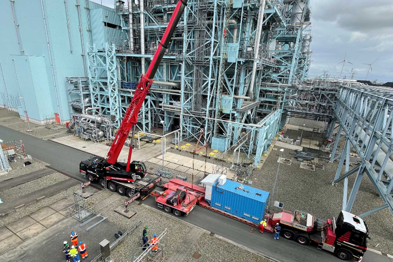 A red crane lifts equipment near a large industrial facility with workers overseeing operations on a gravel path.