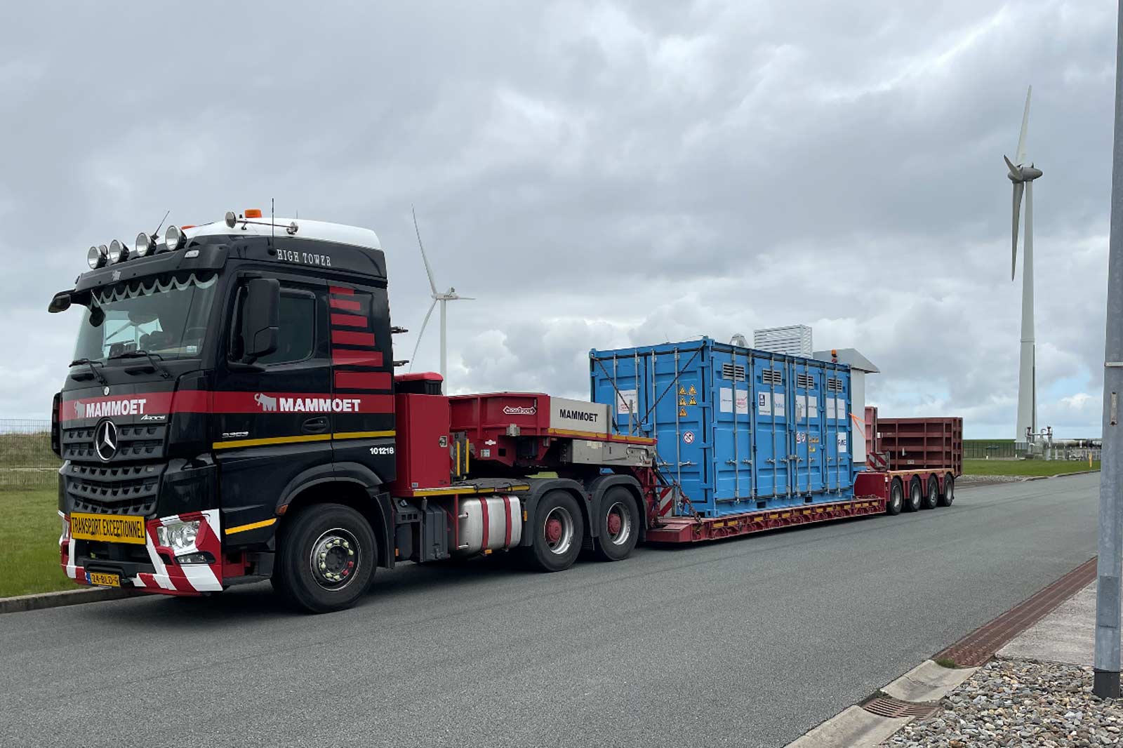 A Mammoet truck transporting a blue container along a road with wind turbines in the background under a cloudy sky.