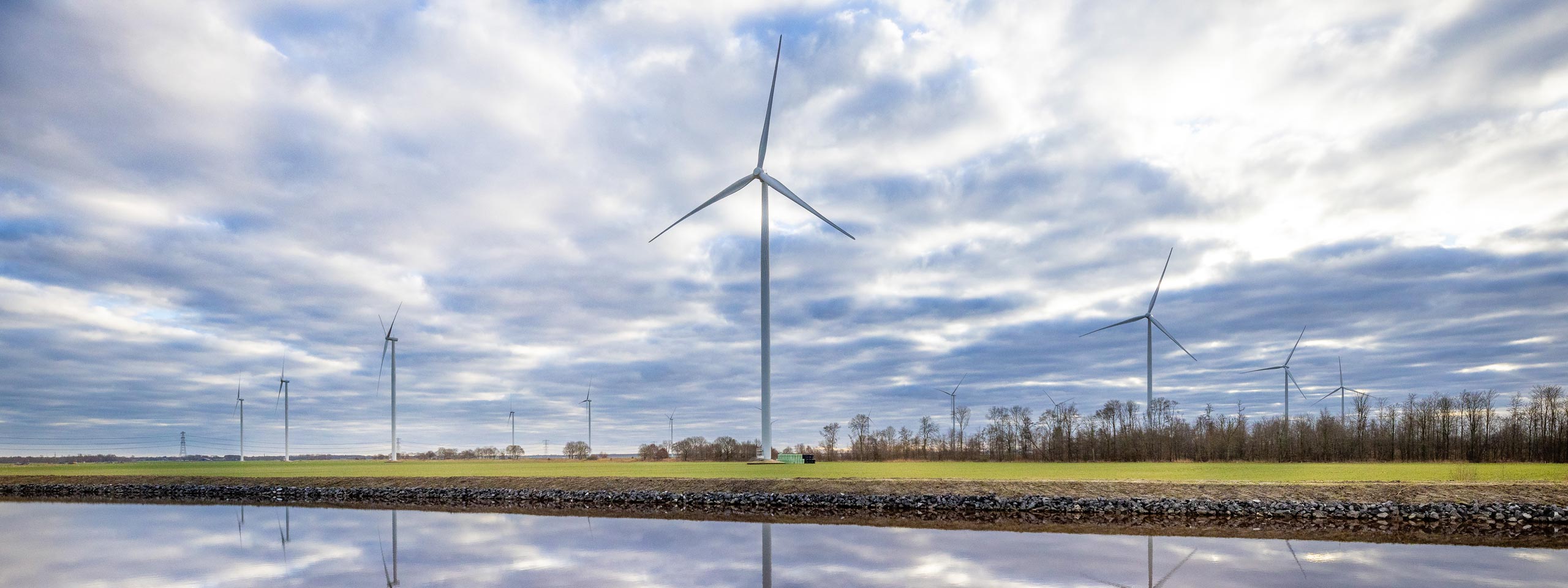 A landscape with several wind turbines against a cloudy sky, reflecting in a calm water body.