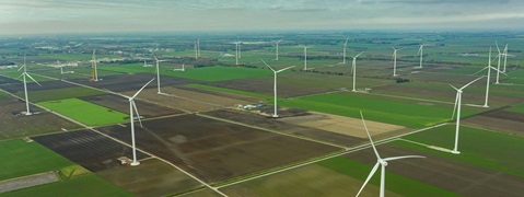 An aerial view of a wind farm with multiple wind turbines set in a patchwork of green and brown fields under a cloudy sky.