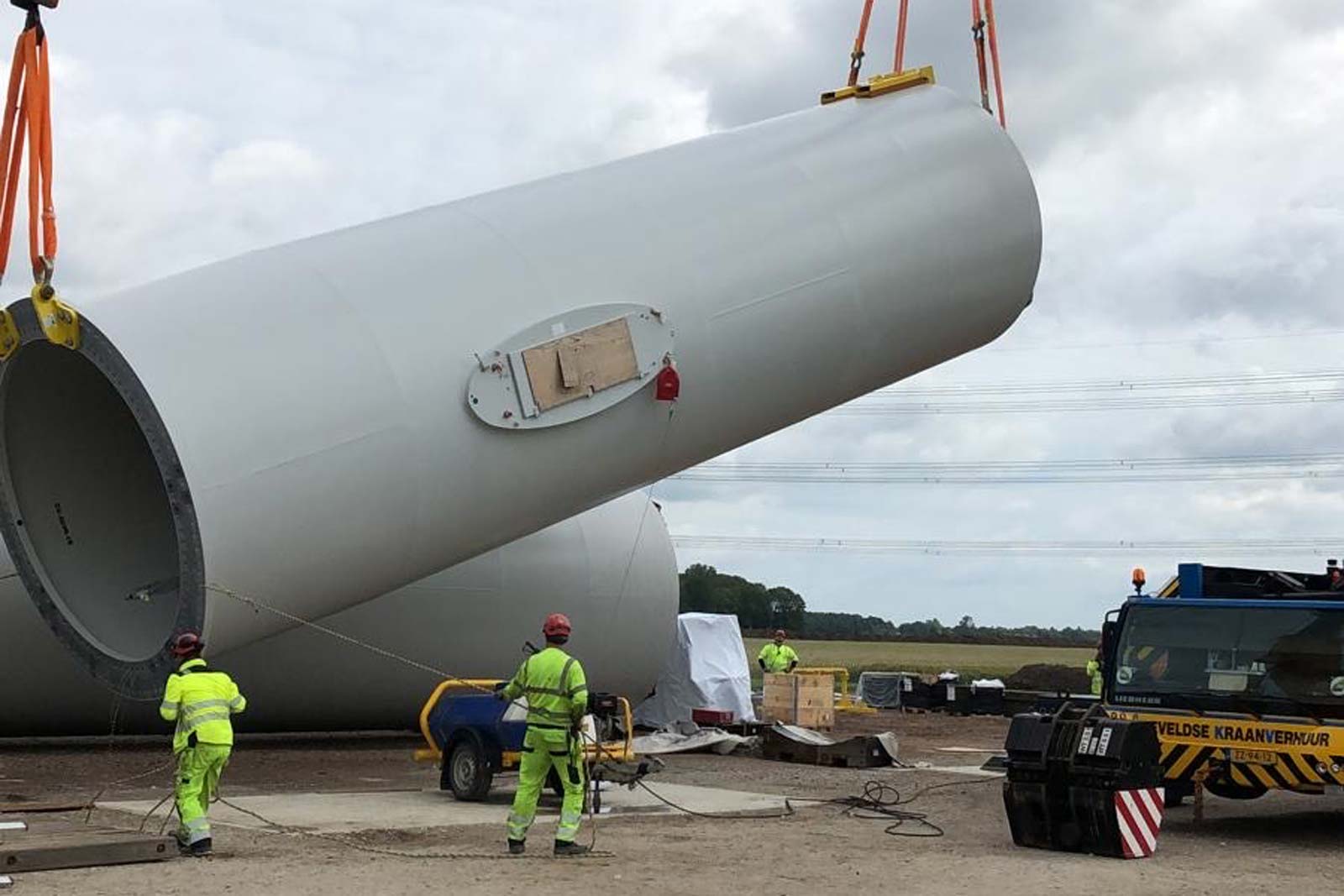 Construction workers in bright clothing are stabilising a large cylindrical wind turbine section being lifted by a crane.