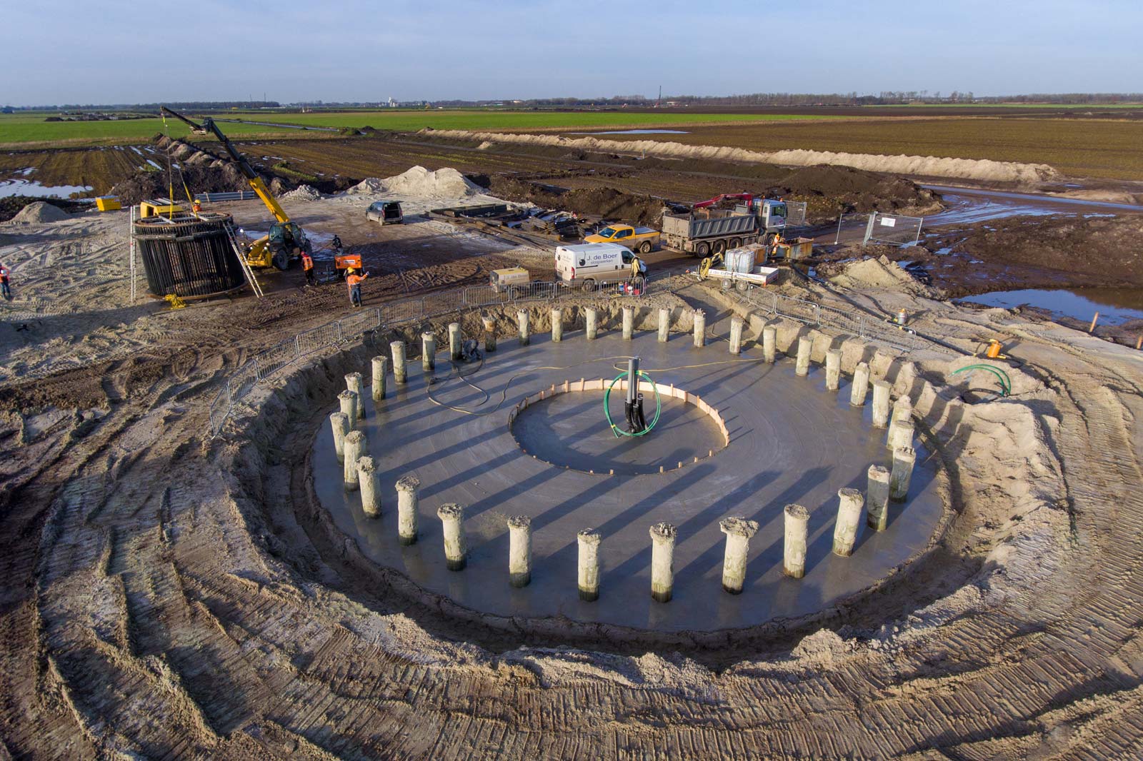 An aerial view of a construction site showing circular foundation pillars and machinery on farmland.