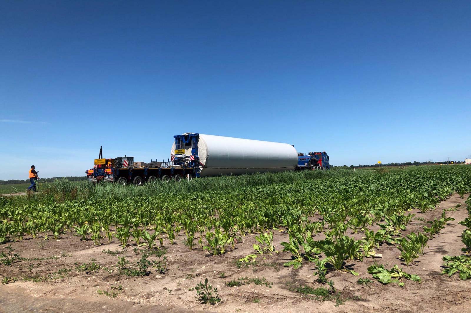A large cylindrical object on a truck being transported through a field, with workers nearby and a clear blue sky above.