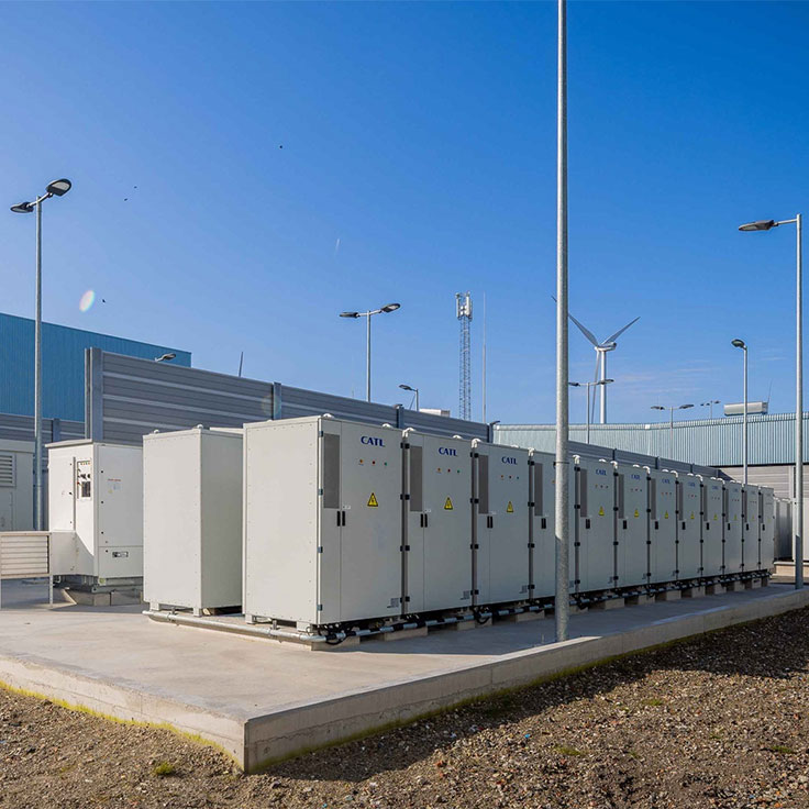 A row of large, grey energy storage containers outside, with a blue sky and wind turbines in the background.