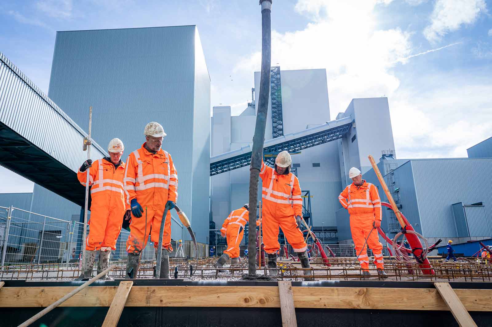 Bouwwerkers in fel oranje uniform werken op een betonplaats met een groot industrieel gebouw op de achtergrond.