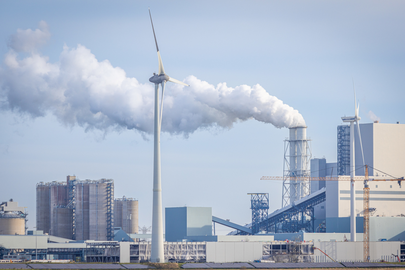 A wind turbine stands adjacent to an industrial facility emitting steam into the sky, set against a clear blue background.