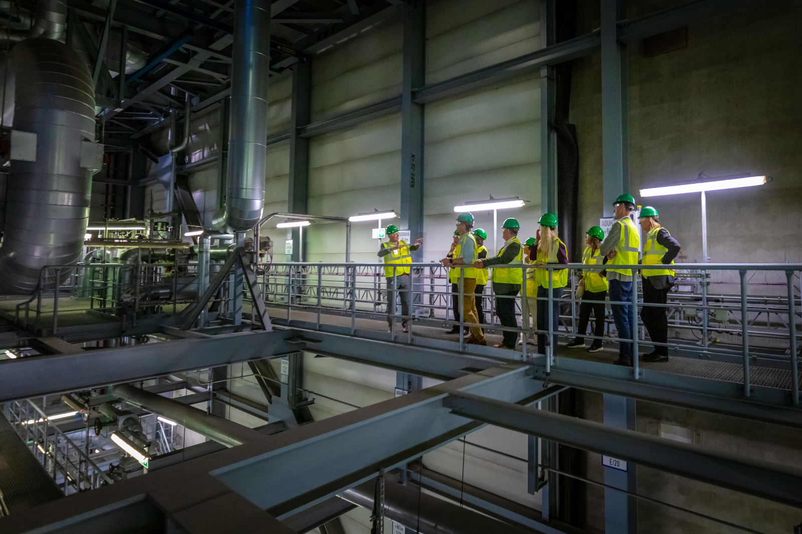 A group of people in safety gear viewing machinery from a walkway in an industrial facility.