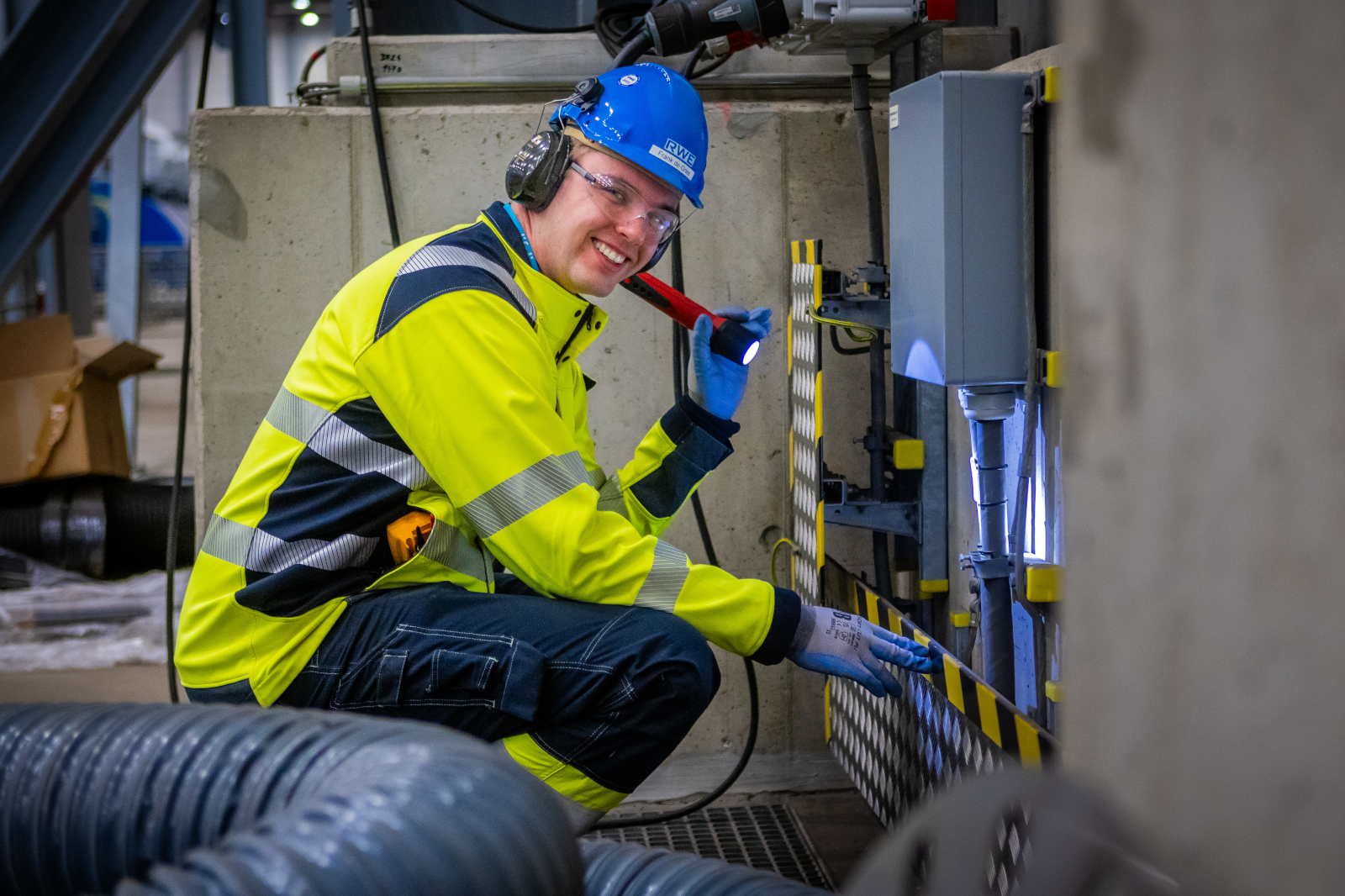 A worker wearing a high-visibility jacket and blue helmet inspects machinery with a flashlight in an industrial setting.