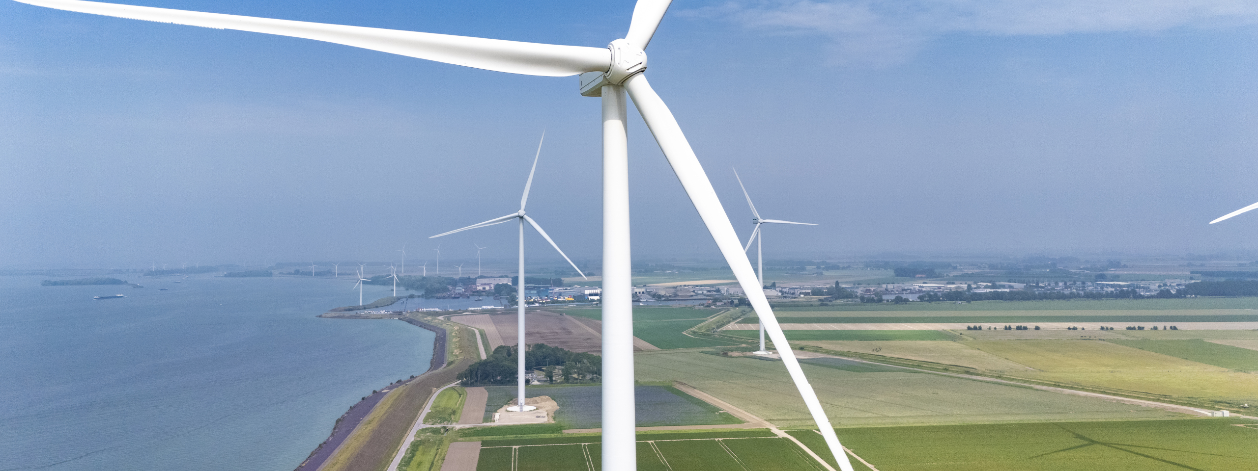 Aerial view of multiple wind turbines near a calm water body, with fields and a rural landscape in the background.