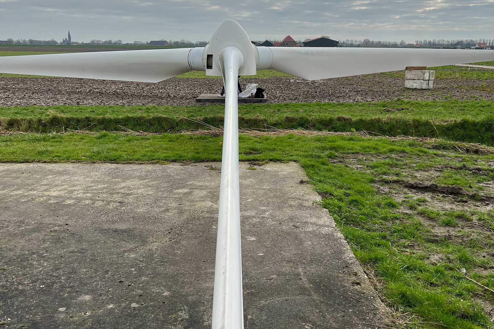 Een close-up van een windturbineblad op een betonnen oppervlak, met velden en een bewolkte lucht op de achtergrond.