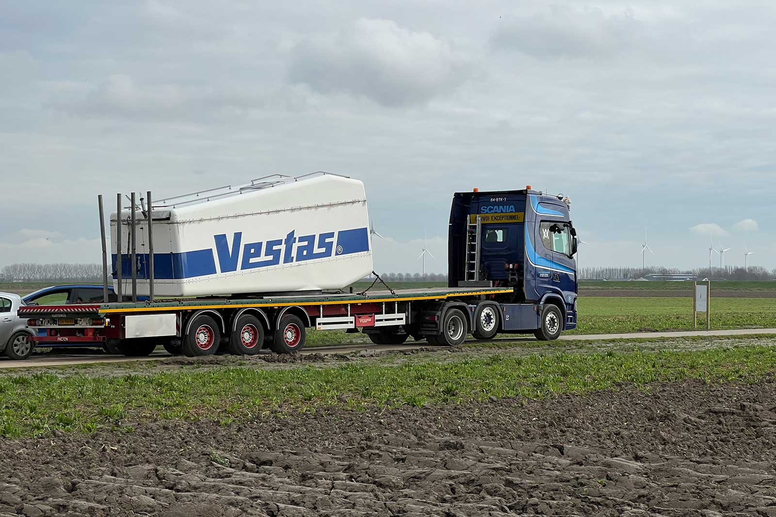 Een blauwe Scania vrachtwagen die een groot Vestas-windturbineonderdeel vervoert langs een landweg, met windturbines op de achtergrond.