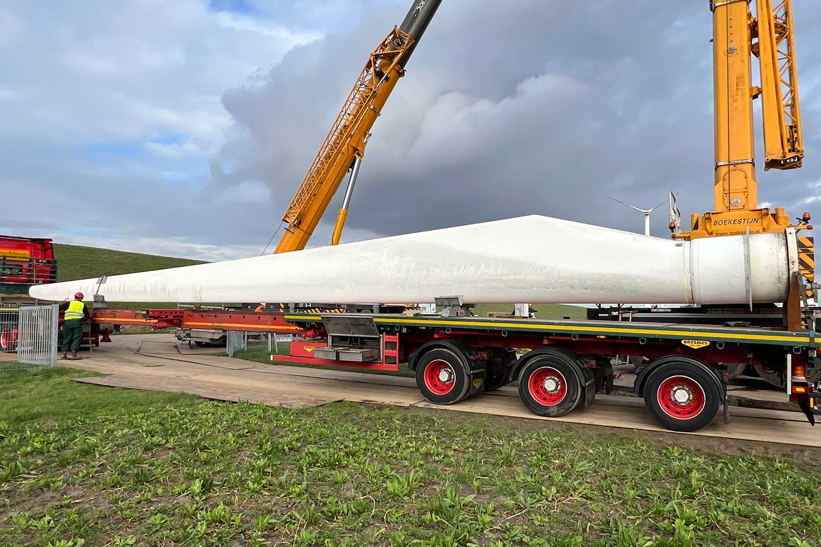 Een kraan heft een groot wit windturbineblad van een vrachtwagen tegen een bewolkte lucht.