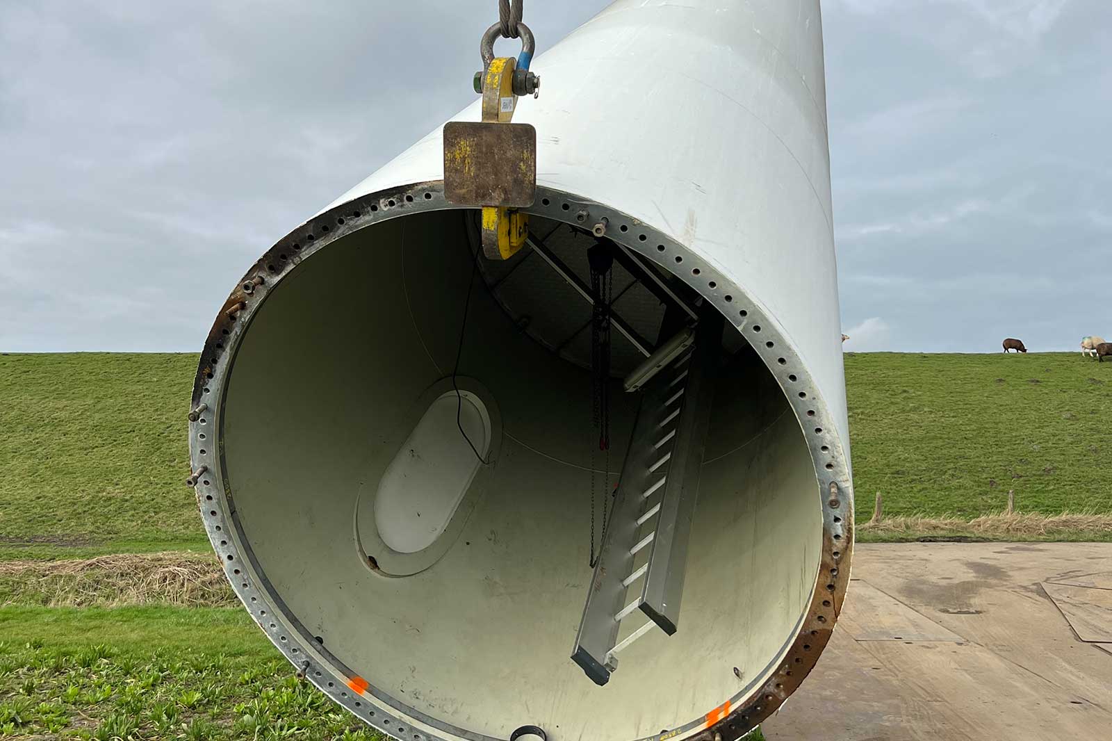 Een grote windturbine toren met een zichtbare ladder binnenin, gehangen door een kraan op een groene achtergrond.