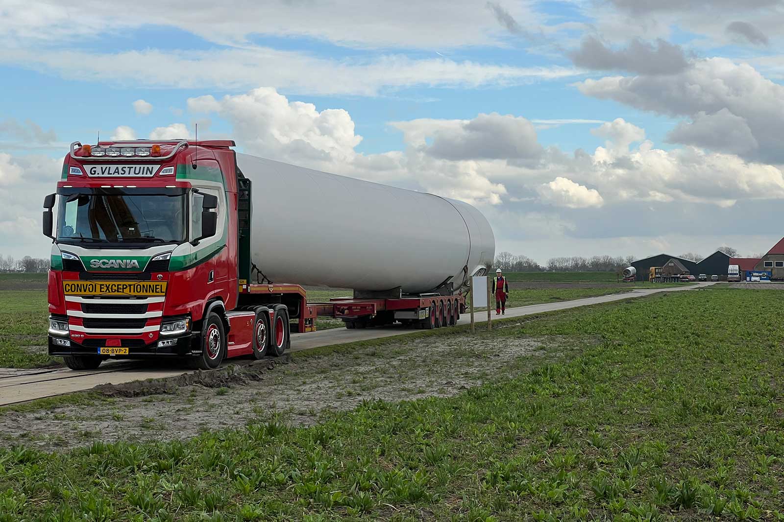 Een Scania-vrachtwagen die een groot wit cilindrisch object vervoert op een dieplader in een landelijke omgeving onder een bewolkte lucht.