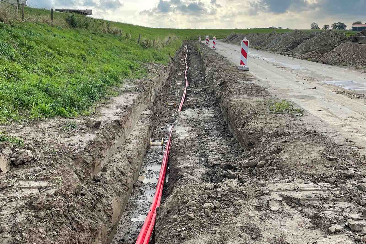 Lange greppel langs weg met rode kabelbuizen en verkeerspalen