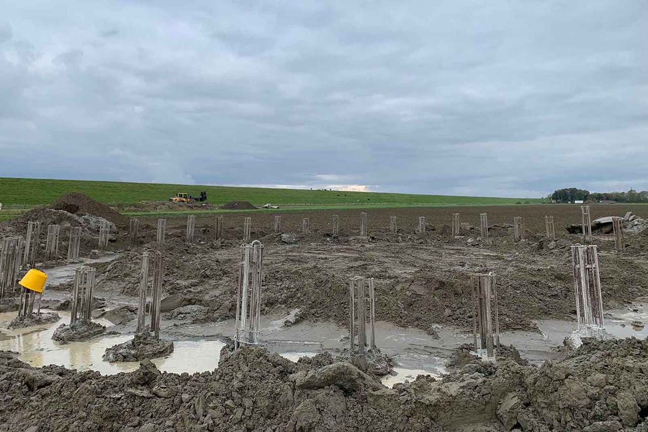 Modderige bouwplaats met betonnen grondpalen met wapeningskooien en gele helm