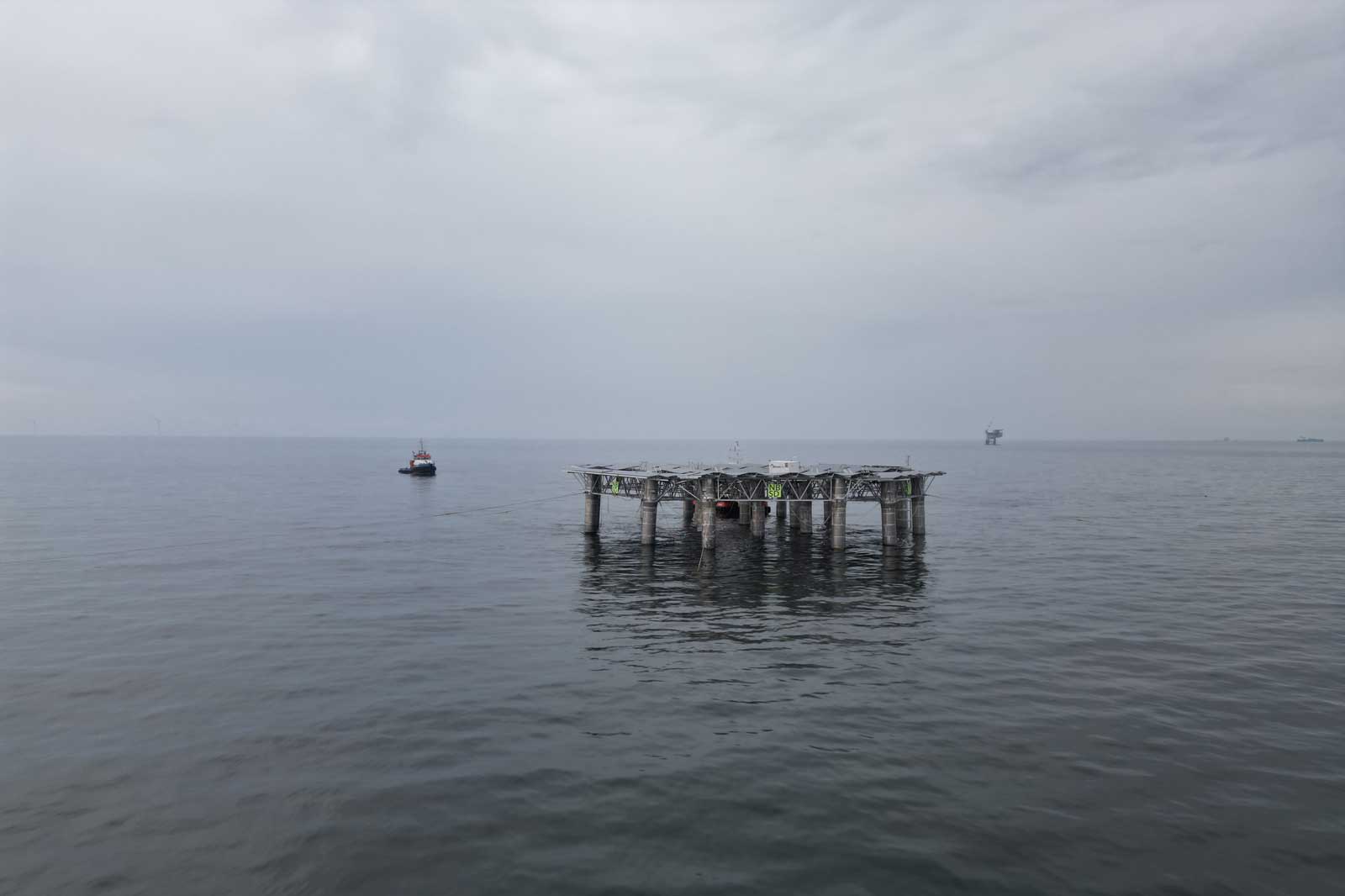 Een zeeplatform staat in kalm water, met een klein bootje in de buurt en verre structuren aan de horizon onder een bewolkte lucht.