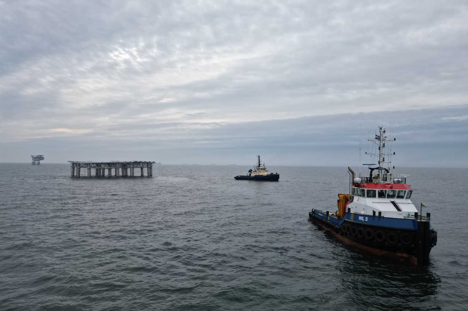 Twee sleepboten navigeren in de zee met een offshore platform op de achtergrond onder een bewolkte lucht.