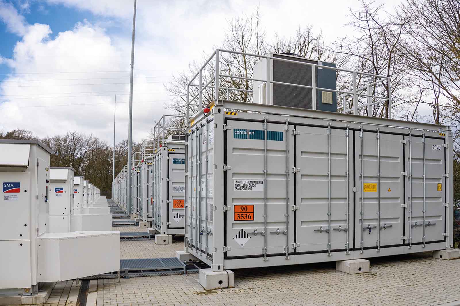 A light-gray industrial equipment container with metal doors and railings sits on a paved yard, flanked by similar units and white cabinets labeled "SMA".