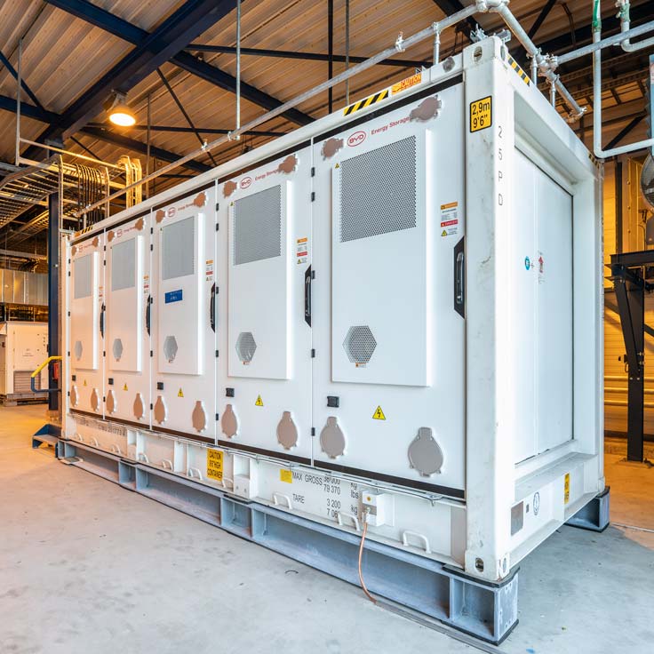 A large white energy storage container with multiple compartments, situated indoors under a metal framework.