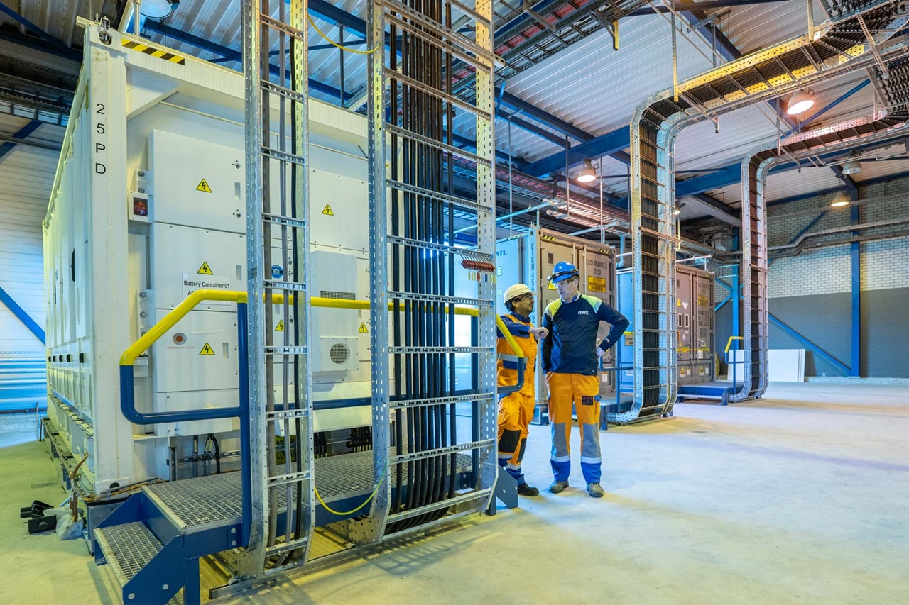 Twee werknemers bespreken naast batterijcontainers in een industriële omgeving, met blauwe stalen balken en veiligheidsborden.