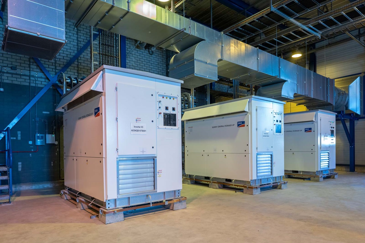 Drie witte omvormers genaamd Sunny Central Storage UP staan in een industriële omgeving met metalen luchtkanalen erboven.