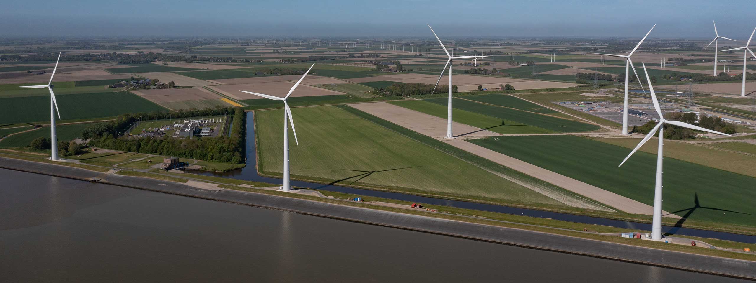 Aerial view of wind turbines in a green landscape, alongside a river with farmlands in the background.