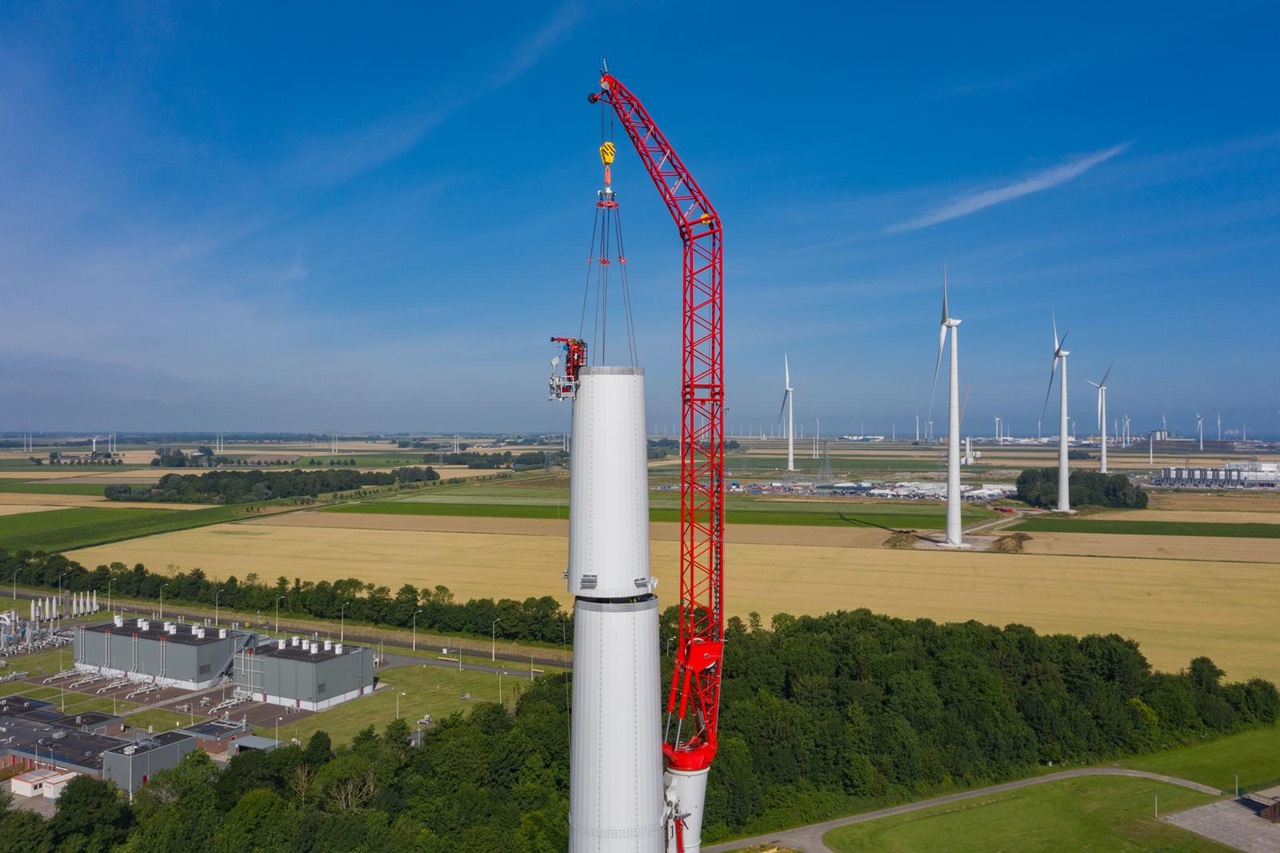 Een windturbine wordt gebouwd, met een rode kraan die onderdelen optilt tegen een blauwe lucht en groene velden.
