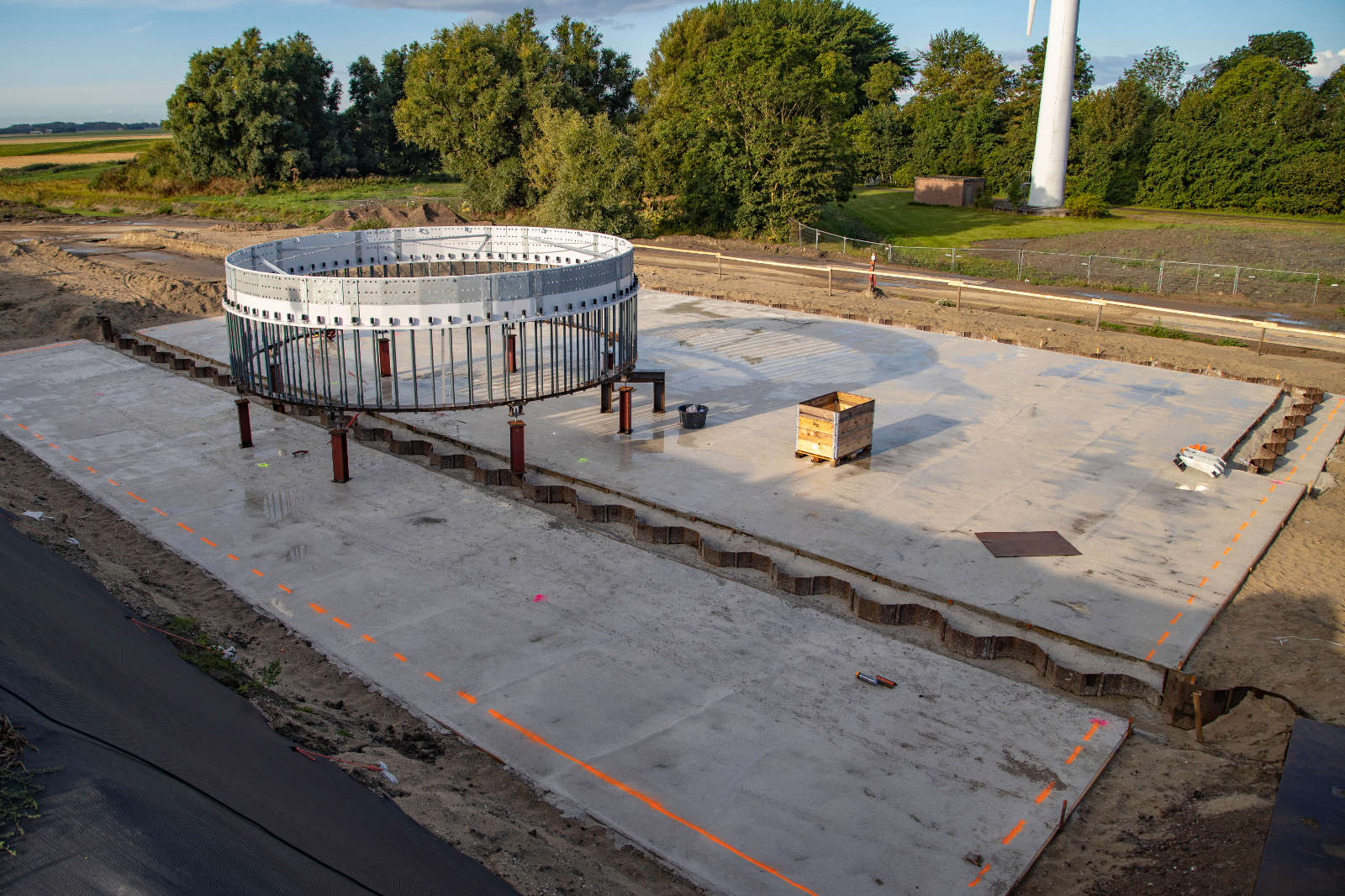 A construction site featuring a circular metal framework on a concrete base, surrounded by trees and machinery.