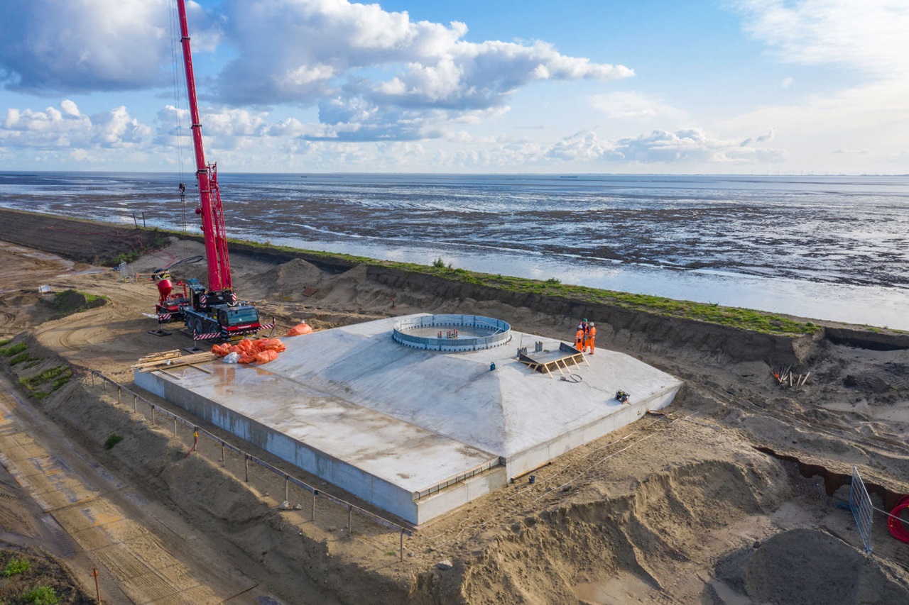 Een bouwplaats met een grote kraan en een betonnen plaat nabij het water. Arbeiders staan op de achtergrond.