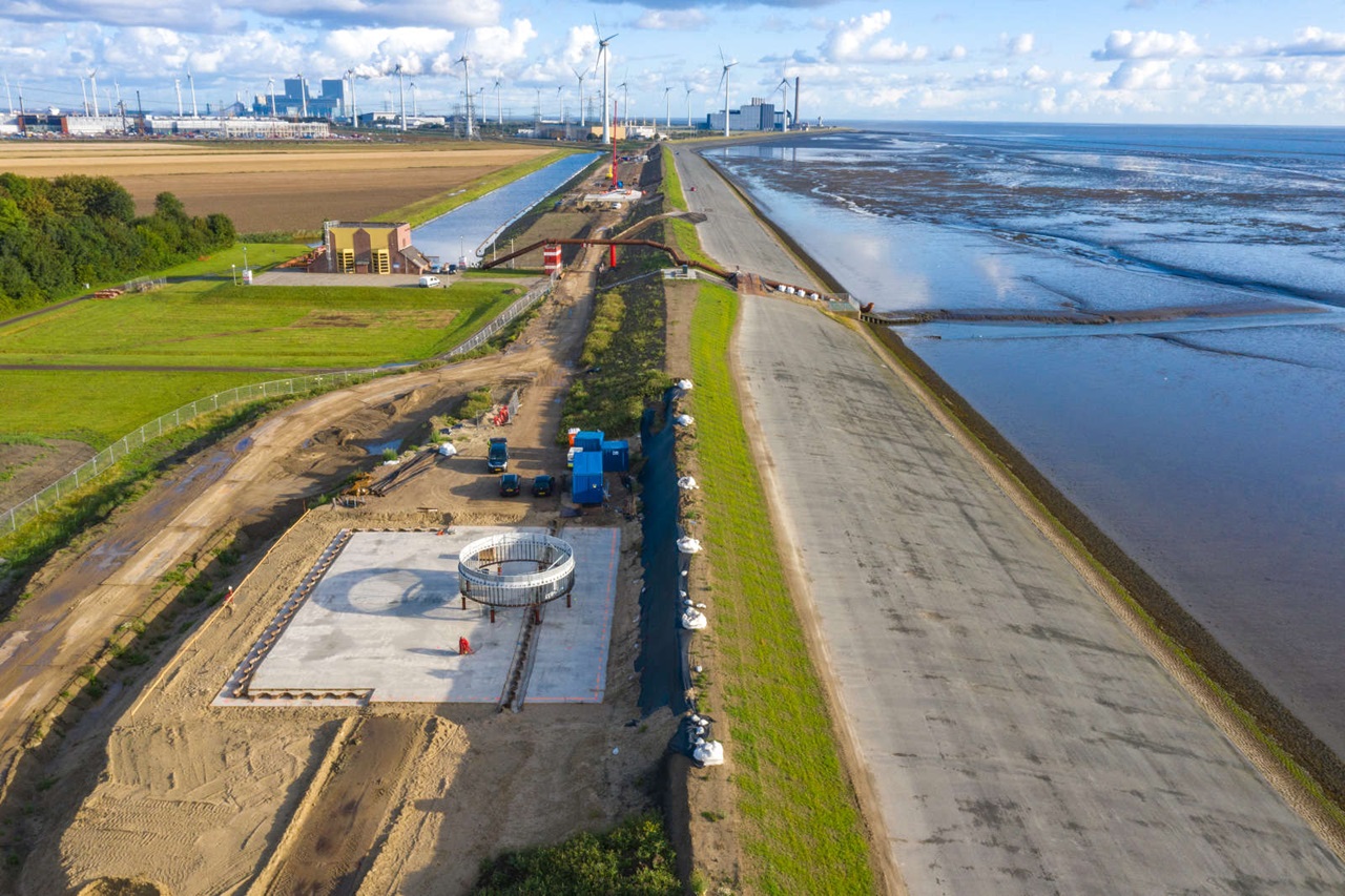 Een bouwplaats met een betonnen platform, een cementring en bouwvoertuigen. Windturbines en een kustgebied zijn op de achtergrond zichtbaar.