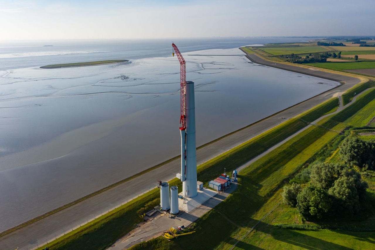 Een hoge windturbine in aanbouw, naast een rustig waterlichaam en groene velden op de achtergrond.